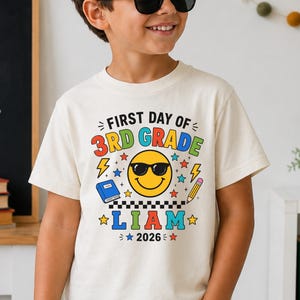 May include: A natural-colored t-shirt featuring a graphic print that reads FIRST DAY OF 3RD GRADE LIAM 2026. The design includes a yellow smiley face wearing sunglasses, a blue book, a yellow pencil, lightning bolts, and colorful stars.