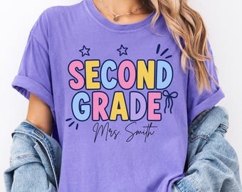 May include: A purple short-sleeve t-shirt featuring the text "SECOND GRADE" in large, colorful block letters, with "Mrs. Smith" written in black cursive script below. The design includes small star and bow graphics in dark blue ink.