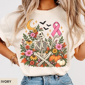 May include: An ivory t-shirt featuring a graphic of a skeleton sitting among colorful flowers, pumpkins, and leaves. The skeleton holds a pink awareness ribbon, with a yellow crescent moon and black bats in the sky above the floral arrangement.