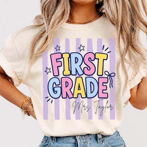 May include: A cream-colored t-shirt featuring a graphic design with vertical light purple stripes. The text reads FIRST GRADE in large, colorful bubble letters, with the name Mrs. Taylor written in black script below, accented by stars.