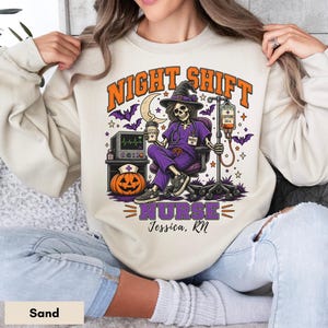 May include: A sand-colored crewneck sweatshirt featuring a graphic of a skeleton nurse in purple scrubs and a witch hat, sitting in a chair with a coffee cup and IV drip. The text reads NIGHT SHIFT NURSE and Jessica, RN, with a pumpkin and monitor.