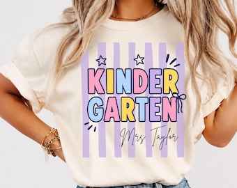 May include: A cream-colored t-shirt featuring a graphic with vertical light purple stripes. The text reads "KINDERGARTEN" in large, colorful block letters, accompanied by "Mrs. Taylor" in black script, two star outlines, and a small bow graphic.