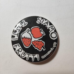 May include: A black and white button with the text "Siamo Tutti Frutti" in a circle around a red apple, a red strawberry, and a bunch of red grapes. The button has a white border and the text "@cosmicolor_pop" in the bottom right corner.