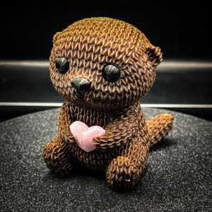 Peut inclure: Figurine d'une loutre brune à la surface texturée, tenant un cœur rose. La loutre a des yeux noirs et un nez noir. La figurine est posée sur une surface sombre.