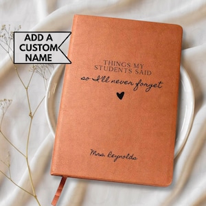 May include: A burnt orange journal with the text "THINGS MY STUDENTS SAID so I'll never forget" and a heart symbol. The name "Mrs. Reynolds" is printed at the bottom. A ribbon bookmark is visible.