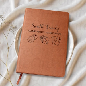 May include: A brown leather-bound game night score book with the text "Smith Family GAME NIGHT SCORE BOOK". The cover features illustrations of dice, playing cards, and dominoes. A brown ribbon bookmark is visible.