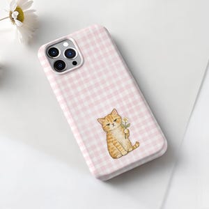 May include: A pink and white gingham patterned phone case featuring a cartoon orange tabby cat holding a daisy. The phone case has a camera cutout and is lying on a white surface.