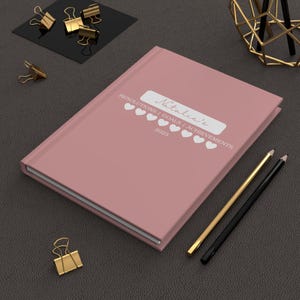 Puede incluir: Un diario rosa con el texto "Natalie's Resolutions | Goals | Achievements 2023" y una fila de corazones.