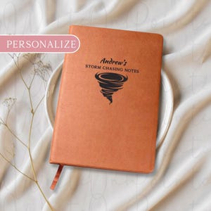 Puede incluir: Un cuaderno naranja personalizado con el texto "Andrew's Storm Chasing Notes" y un gr&aacute;fico de tornado. El cuaderno tiene una cinta de marcador y est&aacute; colocado sobre un plato blanco.