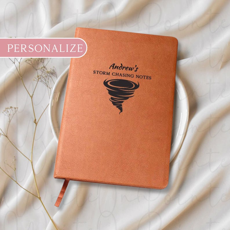 Puede incluir: Un cuaderno naranja personalizado con el texto "Andrew's Storm Chasing Notes" y un gr&aacute;fico de tornado. El cuaderno tiene una cinta de marcador y est&aacute; colocado sobre un plato blanco.