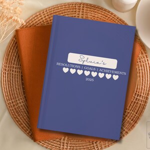 Puede incluir: Un diario de tapa dura azul con el texto "Sylvia's Resolutions | Goals | Achievements 2025" y una fila de corazones blancos.