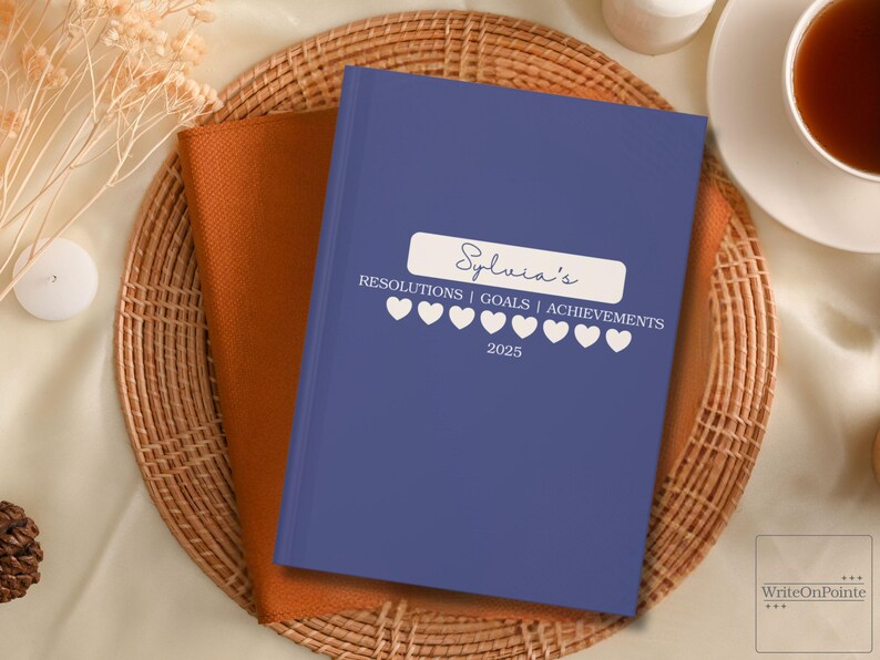 Puede incluir: Un diario de tapa dura azul con el texto "Sylvia's Resolutions | Goals | Achievements 2025" y una fila de corazones blancos.