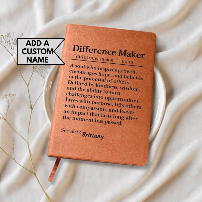 May include: A leather-bound journal in a warm orange hue, featuring the text "Difference Maker" and its definition. The journal includes a ribbon bookmark and is presented on a white plate, with a sprig of delicate greenery.