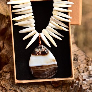 Peut inclure: Un collier avec un pendentif en agate blanc et brun, avec un enroulement de fil de cuivre. Le collier est fait de morceaux de coquille blanche et est présenté dans une boîte avec le texte "60's Daughter by Kristy Cheryl".
