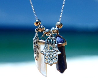 May include: Silver necklace with multiple charms, including a feather, a white pendant, a Hamsa hand, and a dark blue crystal. The necklace has silver beads and a delicate chain. The background is a blurred gradient of blue and green.