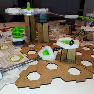 Może przedstawiać: Gra w kulki w kolorze białym i zielonym z drewnianą podstawą i różnymi wieżami i przeszkodami. Gra jest ustawiona na białym stole z szaro-białą planszą sześciokątną.