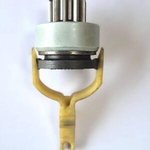 May include: A mechanical starter drive assembly featuring a metallic gear top, a light grey cylindrical housing, a dark grey circular base, and a yellow plastic fork-shaped lever arm extending downwards from the central component.