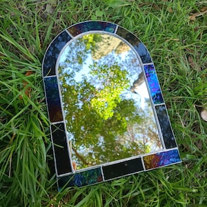 Könnte beinhalten: Ein Spiegel aus Buntglas mit einem schwarzen und schillernden Rahmen. Der Spiegel ist oben gewölbt und spiegelt einen Blick auf Bäume wider.