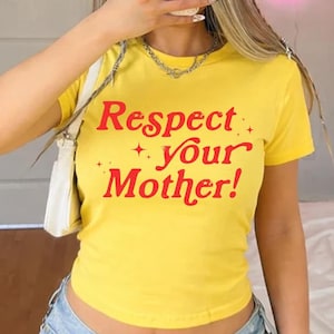 May include: Yellow t-shirt with the text "Respect Your Mother!" in red. The shirt has a crew neck and short sleeves. The text is in a bold, playful font. The shirt is paired with blue denim jeans.