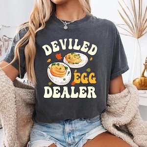 May include: A dark gray t-shirt with the words "DEVILED EGG DEALER" in a retro font. The shirt features a graphic of two deviled eggs on plates. The model is wearing denim shorts and a cream-colored cardigan.