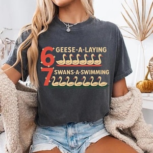 May include: Dark gray t-shirt with the text "6 Geese-A-Laying" and "7 Swans-A-Swimming" in red, yellow, and green. The shirt is worn with denim shorts and a cream-colored cardigan. A decorative pumpkin and woven basket are in the background.