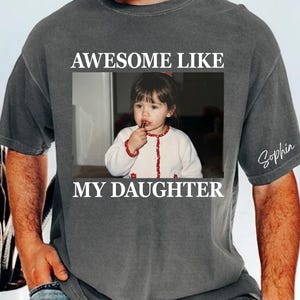 May include: Dark gray t-shirt with a framed photo of a young girl. The text "AWESOME LIKE MY DAUGHTER" is above and below the photo. The name "Sophia" is written on the sleeve.