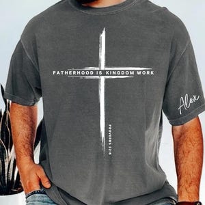 May include: Gray short-sleeve t-shirt with a white cross graphic. The text "FATHERHOOD IS KINGDOM WORK" is above the cross, with "PROVERBS 22:6" below. The name "Alex" is written on the sleeve.