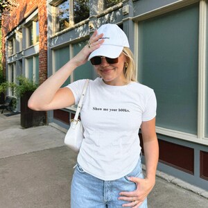 Puede incluir: Camiseta blanca con la frase "Show me your books." y vaqueros azul claro. La persona lleva una gorra de b&eacute;isbol blanca y gafas de sol. Un bolso blanco est&aacute; sobre el hombro.