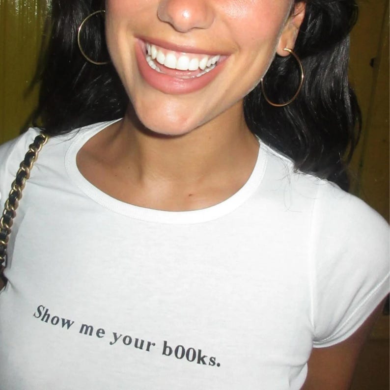 Puede incluir: Camiseta blanca con el texto "Show me your books" en negro. La camiseta es de cuello redondo. La imagen muestra un primer plano de la camiseta y de la persona que la lleva.