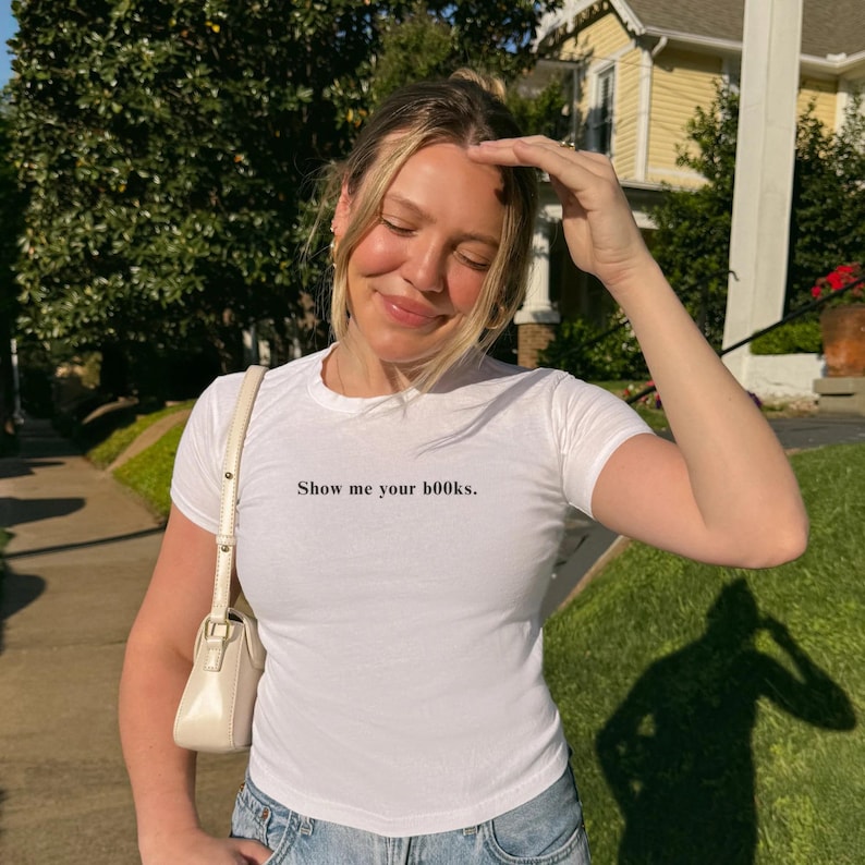 Puede incluir: Camiseta blanca de manga corta con la frase "Show me your books". La camiseta se combina con vaqueros de color azul claro y un bolso de hombro color crema. La foto fue tomada al aire libre en un d&iacute;a soleado.