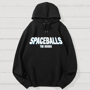May include: Black hoodie with the text "SPACEBALLS THE HOODIE" in white and blue. The hoodie has a drawstring hood and a front pocket. The background is a light wood grain.