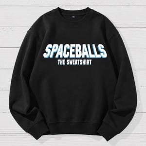 May include: A black long-sleeve crewneck sweatshirt featuring the text SPACEBALLS in large, bold, white and light blue block letters, with the words THE SWEATSHIRT printed in smaller white capital letters directly underneath the main title.