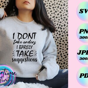 Op de afbeelding: Een grijs sweatshirt met de tekst "I don't take orders I barely take suggestions" in zwart gedrukt. Het sweatshirt wordt gedragen door een persoon met lang donker haar.