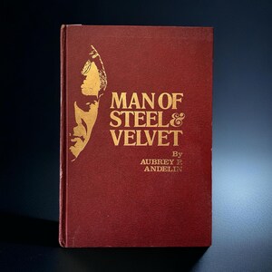 Op de afbeelding: Een boek met een bordeauxrode kaft met de titel "Man of Steel & Velvet" van Aubrey P. Andelin. Het boek heeft gouden letters en een gouden illustratie van een mannengezicht. Het boek staat tegen een donkere achtergrond.