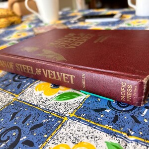 Op de afbeelding: Een bordeauxrode hardcover boek met de titel "Man of Steel & Velvet" met gouden letters. Het boek is licht versleten, met zichtbare slijtage aan de randen. Het boek ligt op een blauw en geel bloemig tafelkleed.
