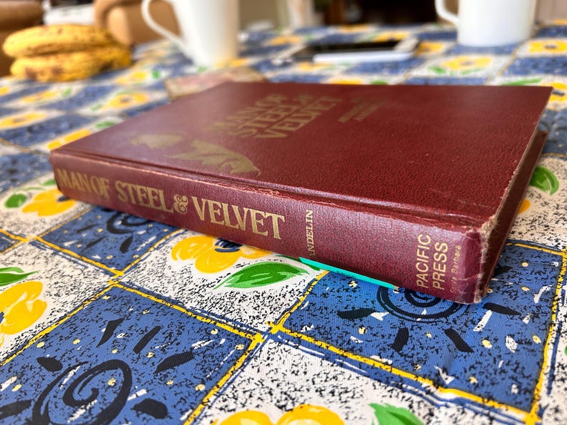 Op de afbeelding: Een bordeauxrode hardcover boek met de titel "Man of Steel & Velvet" met gouden letters. Het boek is licht versleten, met zichtbare slijtage aan de randen. Het boek ligt op een blauw en geel bloemig tafelkleed.