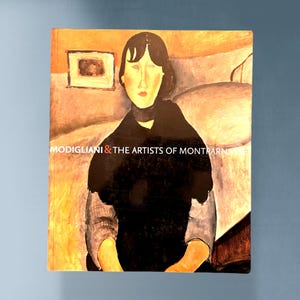 May include: A book cover featuring a Modigliani painting of a woman with dark hair and a black top. The title "MODIGLIANI & THE ARTISTS OF MONTPARNASSE" is printed in white across the bottom of the cover. The background is a warm, muted color.
