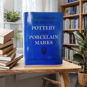 May include: A blue hardcover book titled "Encyclopaedia of British Pottery and Porcelain Marks" by Geoffrey A. Godden. The book is on a wooden table with other books and a bookshelf in the background.