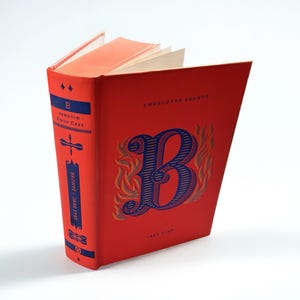 Peut inclure: Un livre rouge intitulé "Jane Eyre" de Charlotte Brontë. La couverture présente une grande lettre "B" bleue avec un design orné. Le dos comporte du texte et un panneau bleu. Le livre est ouvert.