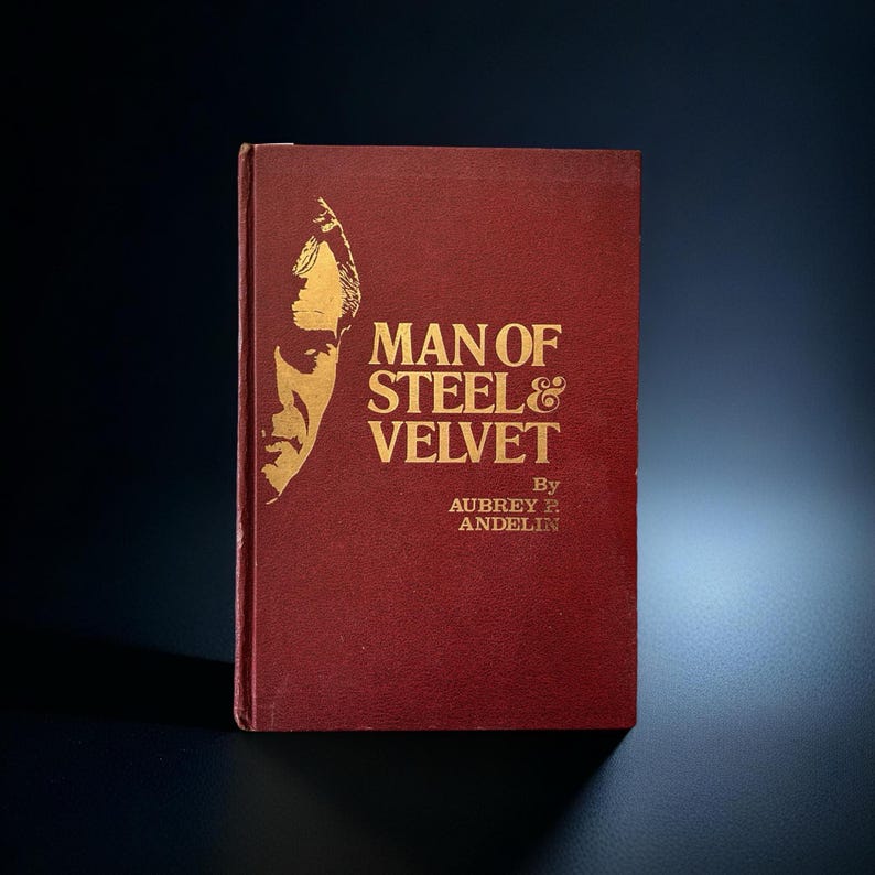 Op de afbeelding: Een boek met een bordeauxrode kaft met de titel "Man of Steel & Velvet" van Aubrey P. Andelin. Het boek heeft gouden letters en een gouden illustratie van een mannengezicht. Het boek staat tegen een donkere achtergrond.