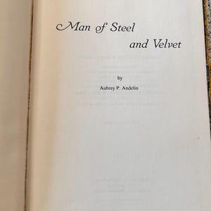 Op de afbeelding: Open boek met de titel "Man of Steel and Velvet" in sierlijke letters. De naam van de auteur, Aubrey P. Andelin, staat eronder. De pagina's zijn gebroken wit met lichtgele randen.