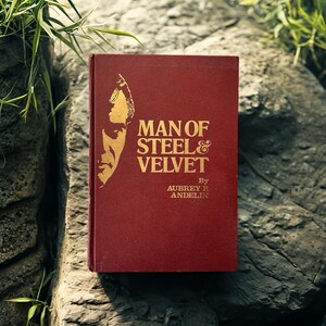 Op de afbeelding: Een rood hardcover boek met de titel "MAN OF STEEL & VELVET" van Aubrey P. Andelin. De titel en een illustratie van een man zijn in goudkleur. Het boek ligt op een grijs stenen oppervlak.