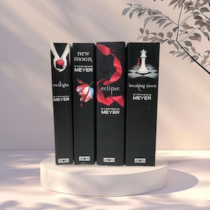Peut inclure: Quatre livres noirs de la saga Twilight de Stephenie Meyer. Les titres des livres sont "Twilight", "New Moon", "Eclipse" et "Breaking Dawn". Chaque livre a une couverture unique avec des accents rouges et blancs. Les livres sont exposés sur un socle circulaire blanc.