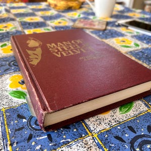 Op de afbeelding: Een bordeauxrode hardcover met de titel "Man of Steel, Velvet" met gouden letters en een portret. Het boek ligt op een tafelkleed met patronen met blauwe, witte en gele vierkanten. De rug van het boek is zichtbaar.