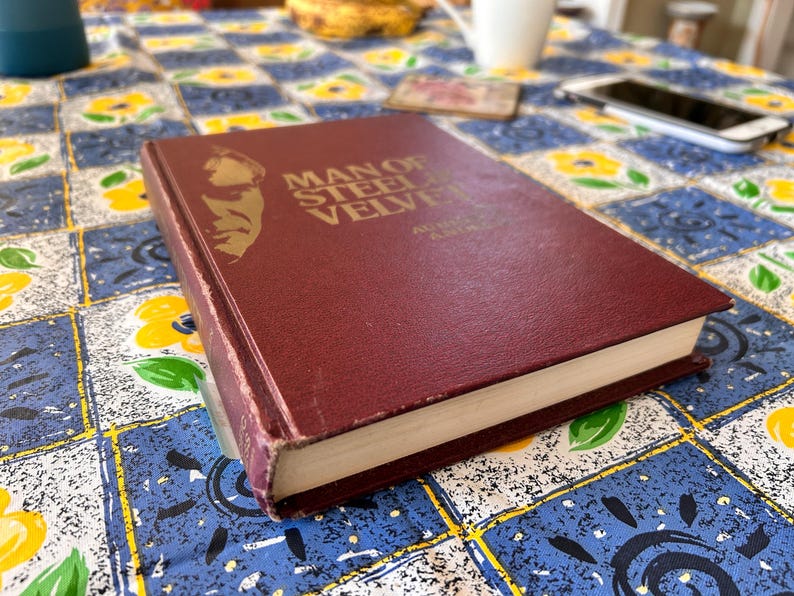 Op de afbeelding: Een bordeauxrode hardcover met de titel "Man of Steel, Velvet" met gouden letters en een portret. Het boek ligt op een tafelkleed met patronen met blauwe, witte en gele vierkanten. De rug van het boek is zichtbaar.