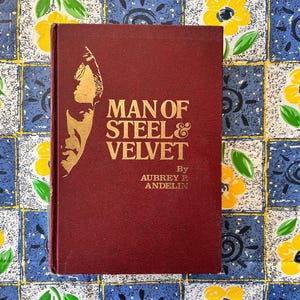 Op de afbeelding: Een bordeauxrode hardcover boek met de titel "MAN OF STEEL & VELVET" van Aubrey R. Andelin. De omslag toont een gouden illustratie van een mannenprofiel. Het boek ligt op een blauw en geel bloemmotief.