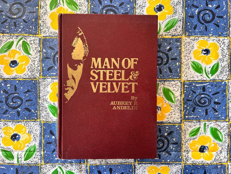 Op de afbeelding: Een bordeauxrode hardcover boek met de titel "MAN OF STEEL & VELVET" van Aubrey R. Andelin. De omslag toont een gouden illustratie van een mannenprofiel. Het boek ligt op een blauw en geel bloemmotief.
