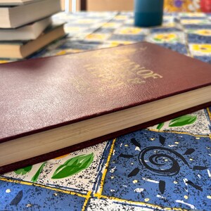 Op de afbeelding: Een bordeauxrood hardcover boek met gouden letters, liggend op een patroon tafelkleed. De pagina's van het boek zijn cr&egrave;mekleurig en andere boeken zijn op de achtergrond gestapeld. Het tafelkleed heeft een blauwe achtergrond met gele en witte bloemmotieven.