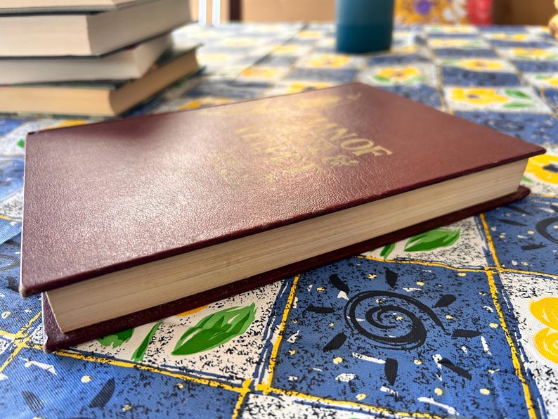 Op de afbeelding: Een bordeauxrood hardcover boek met gouden letters, liggend op een patroon tafelkleed. De pagina's van het boek zijn cr&egrave;mekleurig en andere boeken zijn op de achtergrond gestapeld. Het tafelkleed heeft een blauwe achtergrond met gele en witte bloemmotieven.