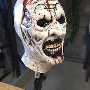 Puede incluir: Una máscara de payaso blanca y grotesca con ojos negros, una sonrisa amplia y amenazante que revela dientes amarillos y vetas rojas similares a sangre. La máscara está montada en un soporte negro, ideal para Halloween.